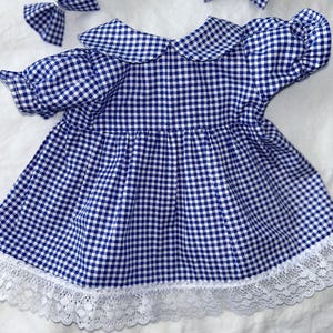Op de afbeelding: Een blauw-wit geruit babyjurkje met witte kanten rand en bijpassende haarstrikken. De jurk heeft een Peter Pan kraag en pofmouwen. De strikken hebben hetzelfde ruitpatroon als de jurk.