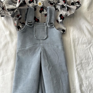 May include: A light blue corduroy overall dress with a front pocket and two buttoned straps, layered over a short-sleeved top featuring a repeating pattern of Mickey Mouse heads and a small white flower-shaped button at the center neckline.
