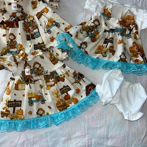 May include: Two white dresses with a fall theme featuring scarecrows, pumpkins, and leaves. The dresses have a blue lace trim and are layered on top of each other. The dress on top has a white collar and short sleeves. The dress on the bottom has long sleeves. There is also a pair of white bloomers with a lace trim.