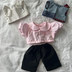 Könnte beinhalten: Eine Sammlung von Puppenkleidung, bestehend aus einem ärmellosen cremefarbenen Oberteil, einem ärmellosen Oberteil mit blau-weißem Vichy-Karo und rotem Paisley-Patchworkmuster, einer hellrosa Kurzarmbluse mit Rundkragen und einer schwarzen Shorts.