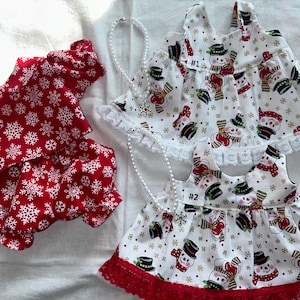 Puede incluir: Dos vestidos de muñeca hechos a mano. Un vestido rojo con copos de nieve blancos y un vestido blanco con estampado de muñeco de nieve. Ambos vestidos tienen ribete de encaje.