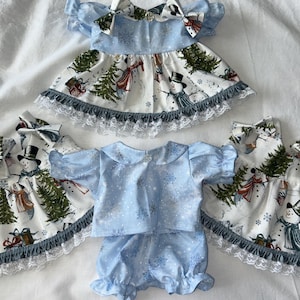 May include: A collection of doll clothing featuring two dresses and a matching top and bloomer set. The garments are made from white fabric with a snowman and pine tree print, and light blue fabric with a white snowflake pattern and lace trim.