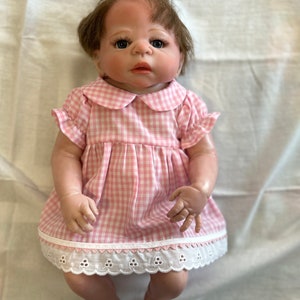 Puede incluir: Un vestido de cuadros vichy rosa y blanco con un ribete de encaje blanco. El vestido tiene un cuello Peter Pan y mangas cortas. La mu&ntilde;eca lleva un lazo rosa en el pelo.