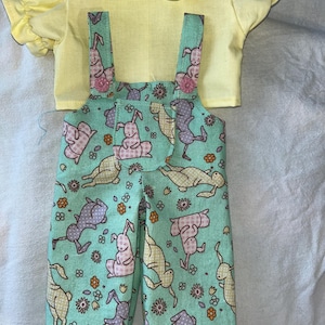 Puede incluir: Un conjunto de muñeca que incluye una camisa amarilla con cuello y mangas con volantes y un peto turquesa. El peto está estampado con conejitos en varias poses, flores y lunares. El conjunto está diseñado para una muñeca.