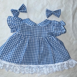 May include: A blue and white gingham dress with a white lace trim and matching hair bows. The dress features a Peter Pan collar and short, puffed sleeves. A traditional, handmade item.