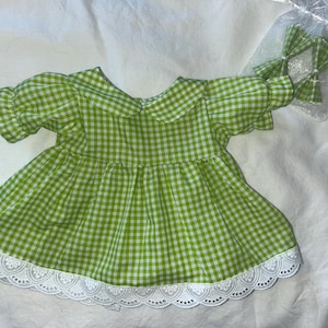 Op de afbeelding: Een jurk met een limegroen en wit vichy-ruitpatroon, korte pofmouwen, een ronde kraag en een witte, geschulpte kanten rand langs de zoom. Inbegrepen is een klein, doorzichtig organza zakje met twee bijpassende groene vichy-ruit haarstrikken.