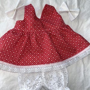 Puede incluir: Un conjunto de vestido y bragas de muñeca para bebé con estampado rojo y blanco. El vestido presenta un estampado de corazones y flores, ribete de encaje blanco y dos lazos blancos. Las bragas son de encaje blanco.