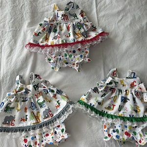 May include: Three sets of sleeveless tops and bloomers featuring a white fabric with a colorful alphabet and animal print. Each top has a ruffled hem with lace trim in red, gray, or green, and the matching bloomers have a multicolored polka dot pattern.