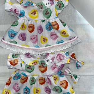 May include: Two white dresses with pink trim and a white lace border. The dresses are covered in colorful candy hearts with various messages, including "Kiss Me", "Be Mine", "Love You", "Sweet", and "Hugs". The dresses are labeled #1 and #2.
