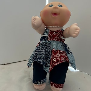 May include: A Cabbage Patch Kid doll wearing a blue and red patchwork dress with a white and blue gingham pattern. The doll has a pearl necklace and is smiling.