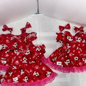 Puede incluir: Dos vestidos rojos con ribete de encaje rosa y estampado de perro. Los vestidos tienen el texto "I woof you" y "Paw". Cada vestido viene con un lazo rosa.
