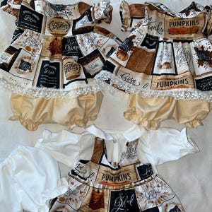 May include: Two matching outfits for baby dolls, each with a brown and black print featuring the words "Give Thanks", "Pumpkins", and "Grateful". Each outfit includes a dress with a lace trim and a pair of beige bloomers. A third outfit with a similar print is also included, featuring a white long-sleeved shirt with a matching dress.