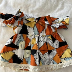 May include: A short-sleeved dress featuring a patchwork pattern of orange, yellow, black, and light blue geometric shapes with black stitched detailing. The dress has an off-the-shoulder neckline with tied sleeves, a single brown button, and a white eyelet lace trim along the bottom hem.