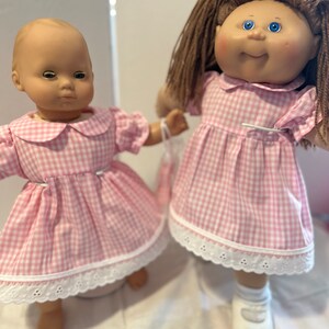 Puede incluir: Dos mu&ntilde;ecas con vestidos de cuadros vichy rosa y blanco con ribete de encaje blanco. La mu&ntilde;eca de la izquierda tiene la cabeza calva y los ojos azules. La mu&ntilde;eca de la derecha tiene el pelo casta&ntilde;o y los ojos azules.