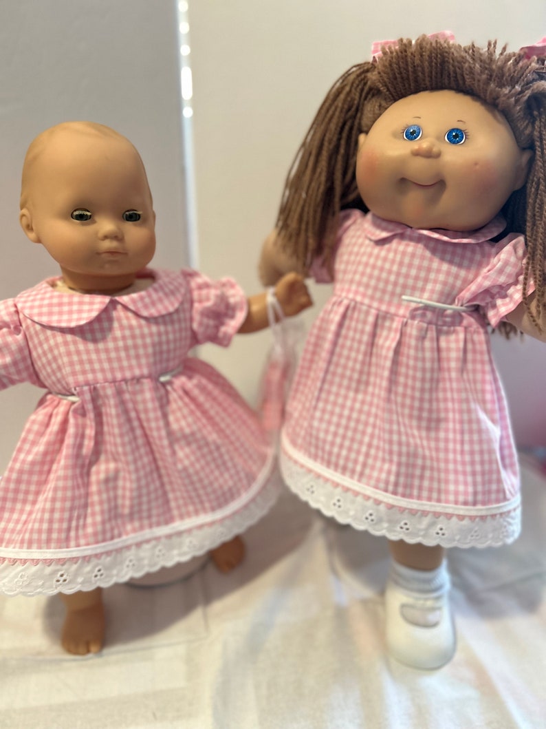 Puede incluir: Dos mu&ntilde;ecas con vestidos de cuadros vichy rosa y blanco con ribete de encaje blanco. La mu&ntilde;eca de la izquierda tiene la cabeza calva y los ojos azules. La mu&ntilde;eca de la derecha tiene el pelo casta&ntilde;o y los ojos azules.