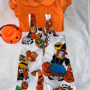 May include: A Halloween costume for a doll, featuring a white and orange jumpsuit with a witch, owl, and pumpkin print, an orange shirt with a Peter Pan collar, and an orange plastic pumpkin.