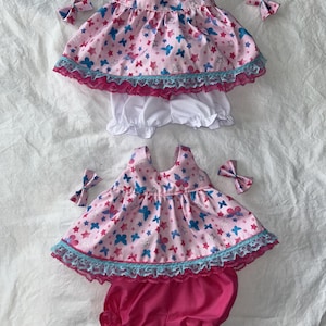 May include: Two doll outfits with matching bows. Each outfit includes a pink top with a butterfly and star pattern, white or pink bloomers, and a matching bow. The tops have blue and pink trim.