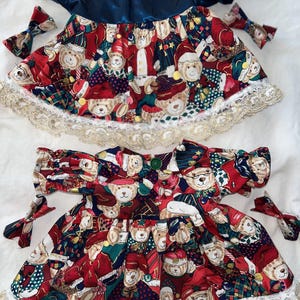 Puede incluir: Dos vestidos con temática navideña con lazos a juego. Los vestidos presentan un corpiño azul marino y una falda con un estampado de osos de peluche con gorros de Papá Noel y atuendos festivos. El dobladillo está adornado con un ribete de encaje dorado.
