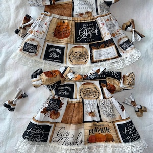 May include: Two white and black dresses with a fall theme. The dresses have a patchwork design with various fall-themed phrases and images, including pumpkins, leaves, and the words "Gather Round", "Give Thanks", and "Farm Fresh Pumpkins". Each dress has a white lace trim and comes with a matching brown and white bow.