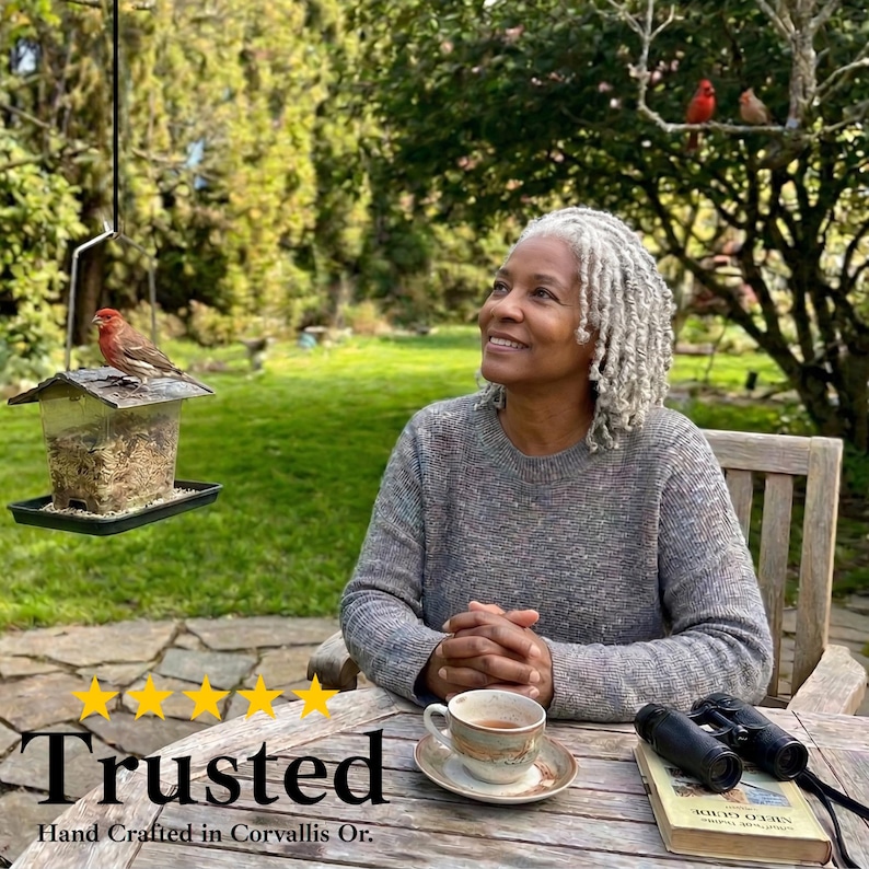 May include: A person with curly gray hair wearing a gray sweater sits at a wooden table with a ceramic teacup, a book, and binoculars. A bird feeder with a small bird hangs nearby, while a red bird is visible in the tree in the background.