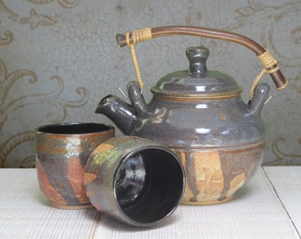 May include: A brown and black ceramic teapot with a wooden handle and two matching cups. The teapot has a lid and a spout. The cups are round and have a wide rim.