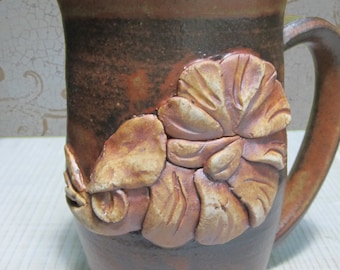 May include: A brown ceramic mug with a raised floral design. The mug has a wide, rounded body and a handle.