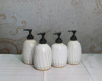 May include: Four ceramic soap dispensers with a white glazed finish and vertical ribbed texture. Each dispenser features a black plastic pump mechanism at the top and a natural clay-colored base, arranged in a group on a white wooden surface.