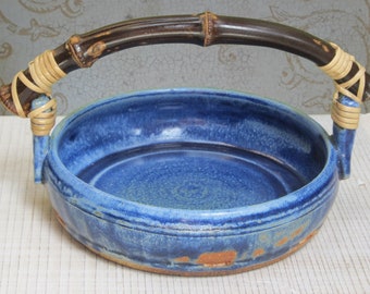 May include: A round, blue ceramic basket with a dark brown bamboo handle. The handle is attached to the basket with woven tan material. The basket has a textured surface and a rustic appearance, suitable for decorative or functional use.