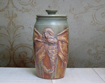 May include: A ceramic jar with a lid, featuring a brown and orange dragon design. The jar has a green glaze and is decorated with a dragon with wings and a tail.