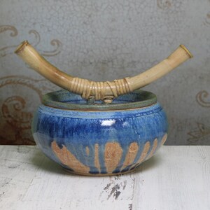 May include: A blue and brown ceramic jar with a lid and a bamboo handle. The jar has a rustic, handmade look.