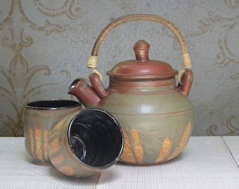 May include: A green and brown ceramic teapot with a woven handle and two matching ceramic cups. The teapot has a lid and a spout. The cups have a black interior.