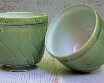 May include: Two ceramic bowls with a glossy light green glaze. The exterior of each bowl features a diagonal ribbed texture and a horizontal band near the rim decorated with small, stamped leaf-like patterns. One bowl stands upright while the other is tilted.