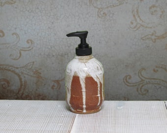 May include: A ceramic soap dispenser with a rounded body featuring a textured, reddish-brown clay base and a white glaze that drips down the sides. The dispenser is topped with a functional black plastic pump mechanism for dispensing liquid soap.