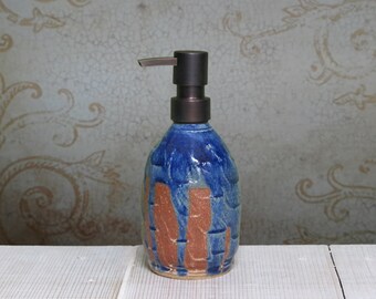 May include: A ceramic soap dispenser with a rounded body featuring a textured, mottled blue glaze over a raw, unglazed reddish-brown clay base. The dispenser is topped with a dark bronze-colored metal pump mechanism for dispensing liquid soap.