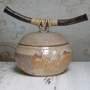 May include: A ceramic jar with a lid and a wooden handle wrapped in woven straw. The jar is glazed in shades of brown, beige, and white.