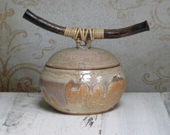 May include: A ceramic jar with a lid and a wooden handle wrapped in woven straw. The jar is glazed in shades of brown, beige, and white.