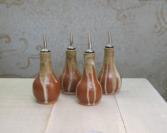 May include: Four ceramic oil bottles with stainless steel spouts. The bottles are a warm brown with vertical cream-colored stripes and a dripped glaze effect. They are arranged on a light-colored wooden surface.