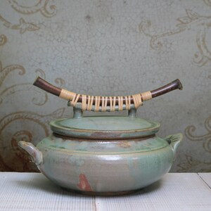 May include: A green ceramic pot with a lid and a wooden handle wrapped in woven straw. The pot has a rustic, handmade look.