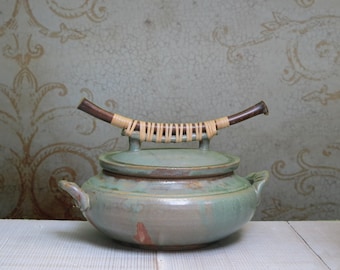 May include: A green ceramic pot with a lid and a wooden handle wrapped in woven straw. The pot has a rustic, handmade look.
