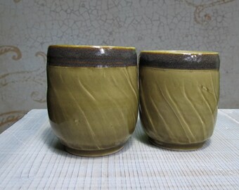 May include: Two ceramic cups featuring a glossy olive green glaze with a subtle diagonal ribbed texture on the body and a dark brown band around the rim. The cups have a rounded shape and a smooth finish, showcasing a consistent design across both pieces.