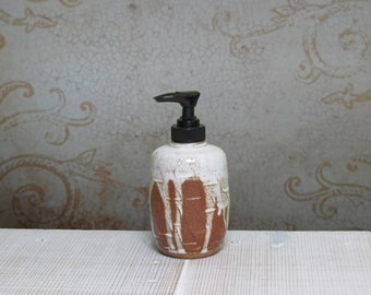 May include: A ceramic soap dispenser with a cylindrical body featuring a textured, reddish-brown clay base partially covered by a thick, white, drippy glaze. The dispenser is topped with a functional black plastic pump mechanism for dispensing liquid.