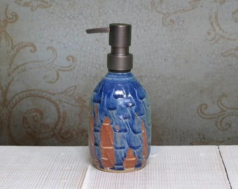 May include: A ceramic soap dispenser with a rounded body featuring a textured, overlapping scale pattern in shades of deep blue and exposed terracotta brown. The vessel is topped with a dark metallic pump mechanism for dispensing liquid soap.