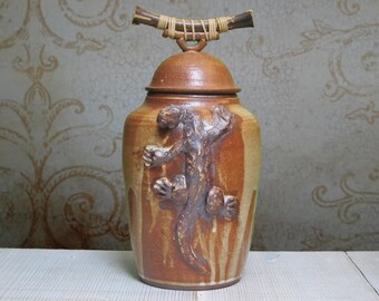 May include: A handcrafted ceramic jar with a lid, featuring a detailed lizard sculpture on the front. The jar is a warm brown color with a textured surface and a woven handle on top. The jar is approximately 10 inches tall.