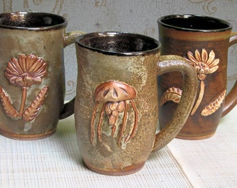 May include: Three ceramic mugs with different designs. The first mug has a brown and green glaze with a raised flower design. The second mug has a brown and green glaze with a raised jellyfish design. The third mug has a brown and green glaze with a raised flower design.