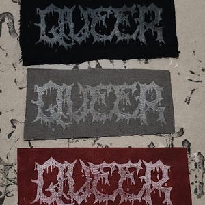 Könnte beinhalten: Drei rechteckige Aufnäher mit dem Wort "QUEER" in einer tropfenden, gotischen Schriftart. Die Aufnäher sind schwarz, grau und rot. Der Text ist in einer kontrastierenden Silberfarbe gehalten. Die Aufnäher sind wahrscheinlich für Kleidung oder Accessoires bestimmt.