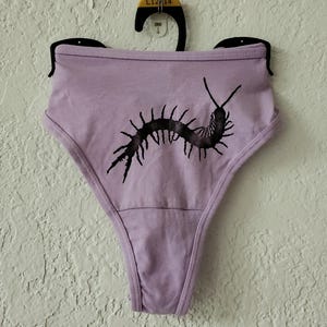 May include: Lavender high-waisted underwear with a black centipede graphic. The underwear is hanging on a black hanger against a textured white wall. The centipede design is centered on the front.
