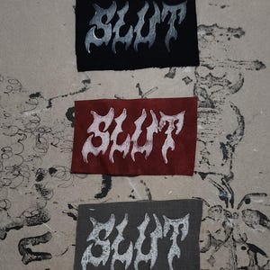 May include: Three rectangular patches in black, red, and gray, each bearing the word "SLUT" in a stylized, white, flame-like font. The patches are arranged vertically against a textured, off-white background.