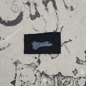 May include: A small, rectangular black patch with a light gray, abstract design in the center. The patch is set against a textured, off-white background covered in dark gray markings and scribbles.