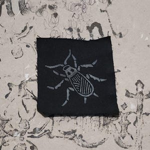 Puede incluir: Un parche de tela negra cuadrada con un diseño de insecto gris claro. El insecto tiene seis patas y un cuerpo segmentado. El parche tiene un borde ligeramente deshilachado. El fondo es una superficie texturizada blanquecina con marcas gris oscuro.