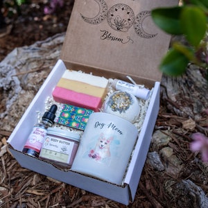 May include: A gift box with a variety of self-care items. The box includes a bar of soap, a bath bomb, a candle with a dog graphic, body oil, body butter, and a lip balm. The box is labeled "The Divine Blossom".